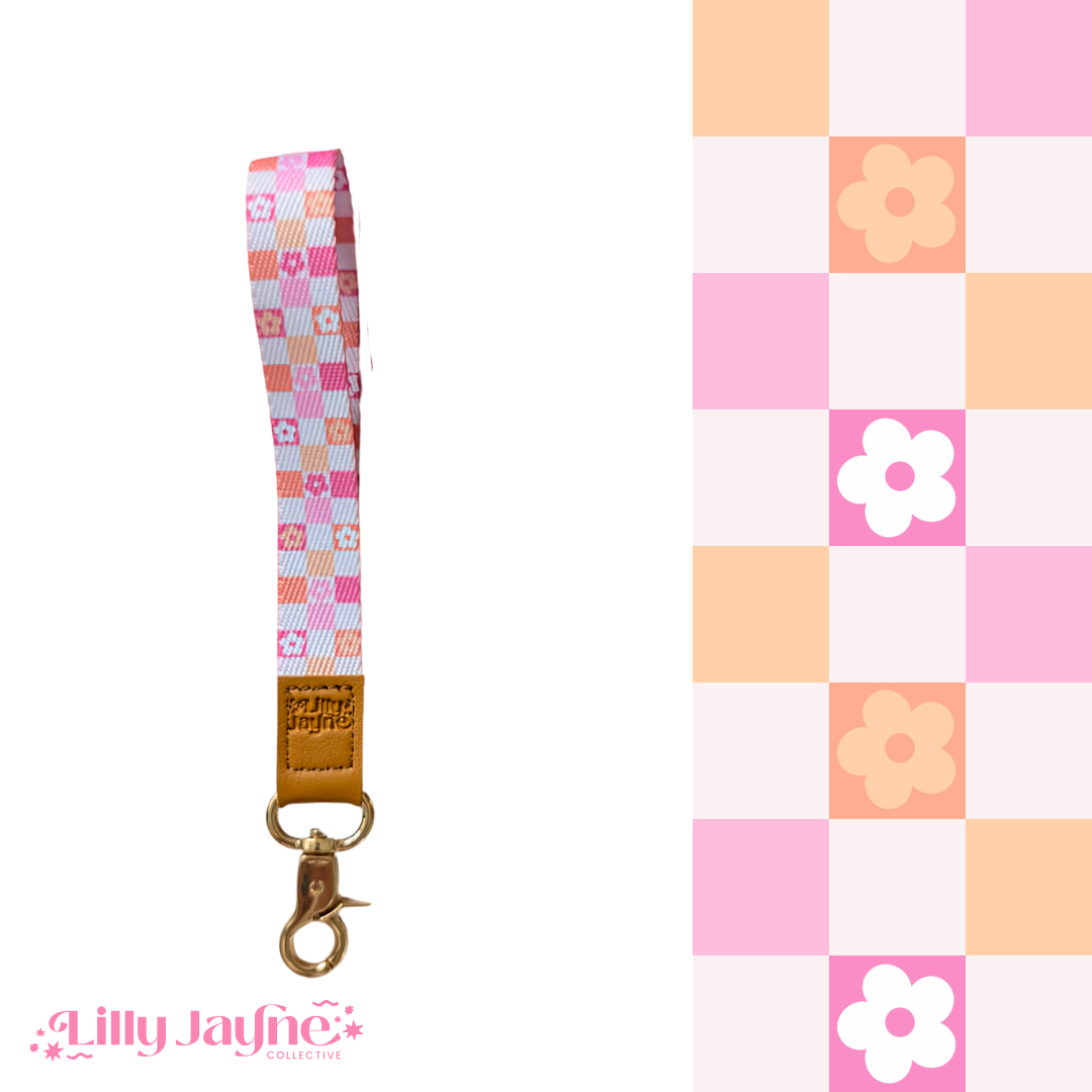 Three colourful fabric wristlet keychains with gold hardware and tan leather ends, featuring pink gingham daisy, blush polka dot stripe, and retro orange checker floral patterns, laid on pastel notebooks with floral accents – Lilly Jayne Collective.