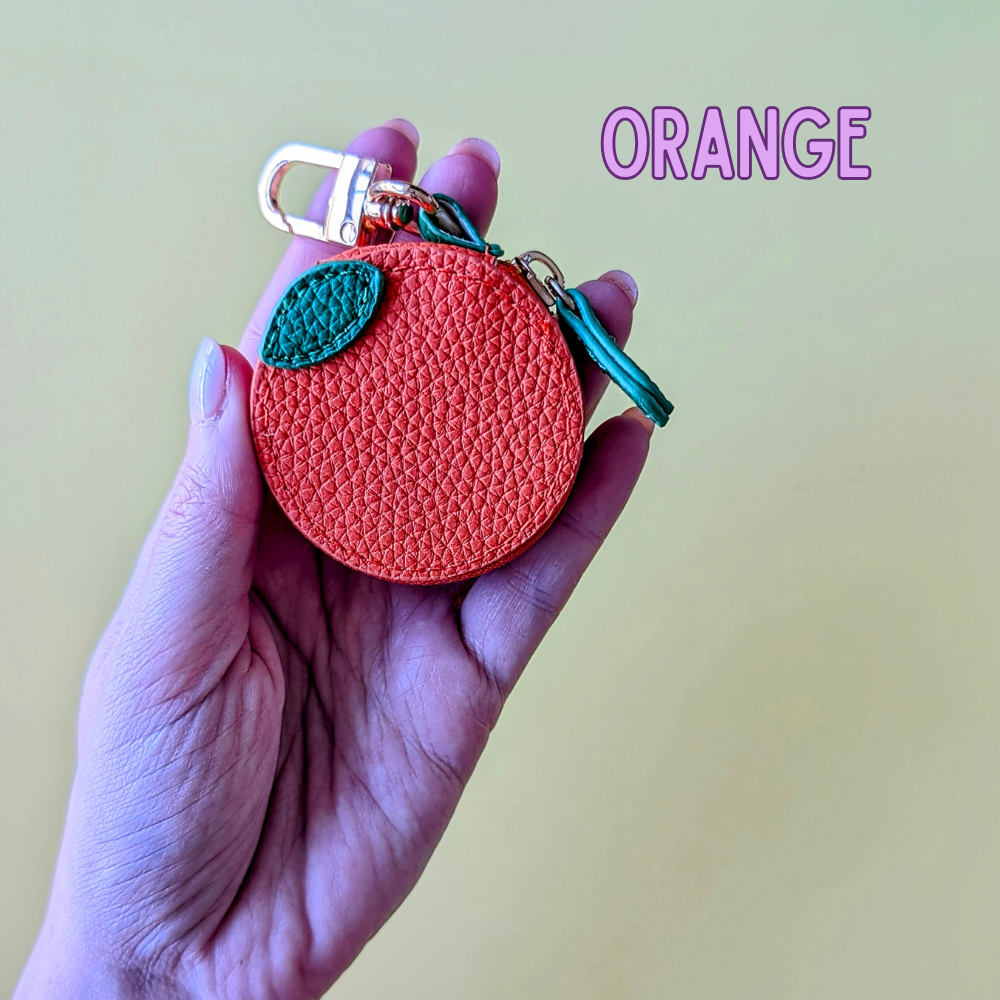 Hand holding an orange-shaped keychain with a green leaf on a light yellow background.