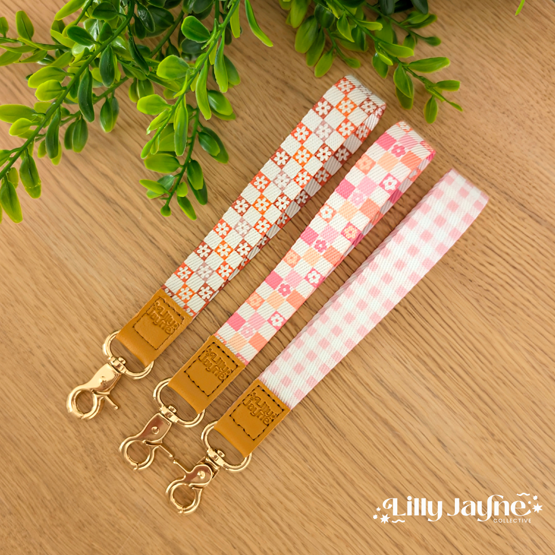 Three colourful fabric wristlet keychains with gold hardware and tan leather ends, featuring pink gingham daisy, blush polka dot stripe, and retro orange checker floral patterns, laid on pastel notebooks with floral accents – Lilly Jayne Collective.