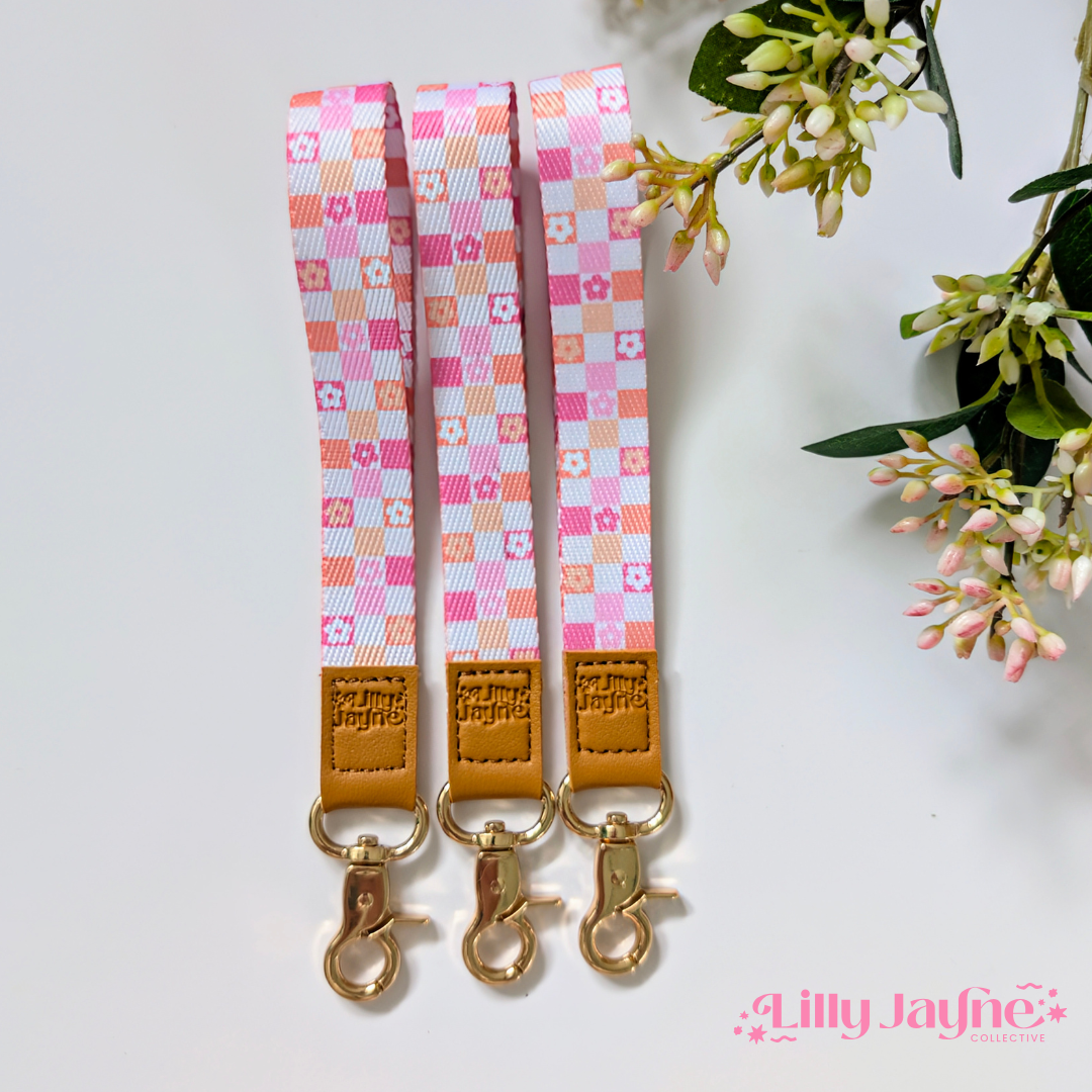 Three colourful fabric wristlet keychains with gold hardware and tan leather ends, featuring pink gingham daisy, blush polka dot stripe, and retro orange checker floral patterns, laid on pastel notebooks with floral accents – Lilly Jayne Collective.