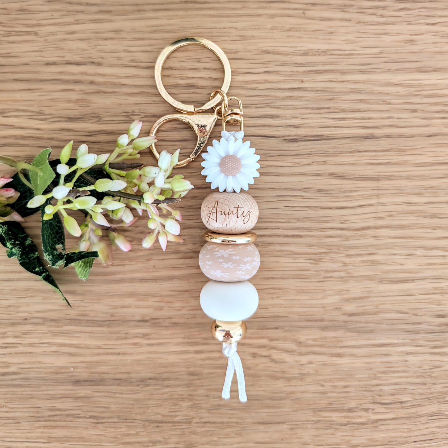 Handmade keyring with white daisy flower charm, engraved 'Aunty' wooden bead, floral patterned bead, and pastel pink accents on a wooden surface with greenery
