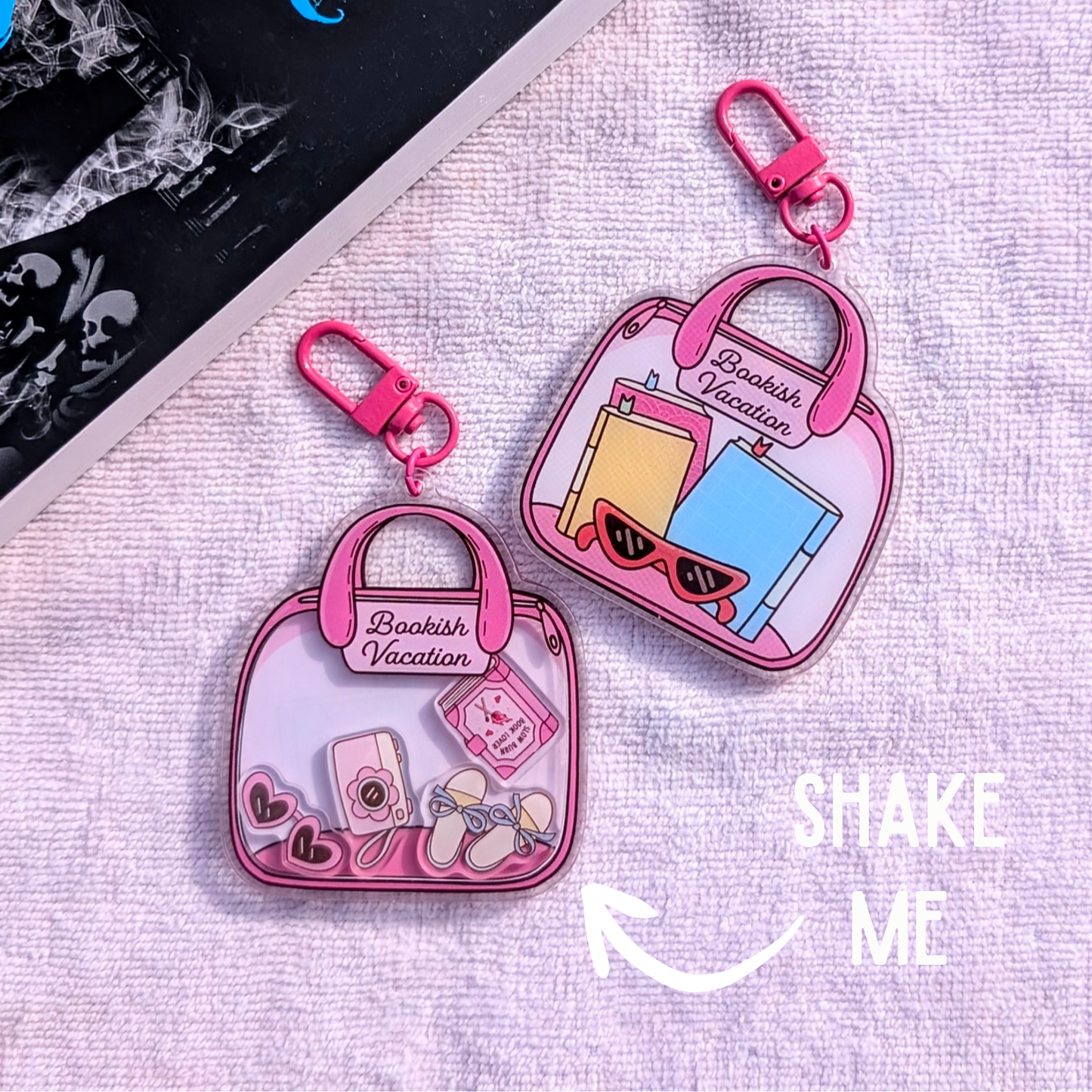 Pink 'Bookish Vacation' keychains on a textured surface with 'Shake Me' text.