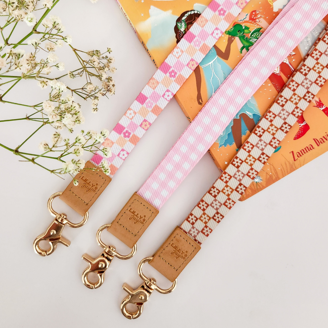 Three colourful fabric wristlet keychains with gold hardware and tan leather ends, featuring pink gingham daisy, blush polka dot stripe, and retro orange checker floral patterns, laid on pastel notebooks with floral accents – Lilly Jayne Collective.