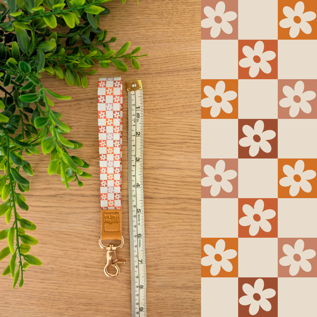 Three colourful fabric wristlet keychains with gold hardware and tan leather ends, featuring pink gingham daisy, blush polka dot stripe, and retro orange checker floral patterns, laid on pastel notebooks with floral accents β Lilly Jayne Collective.