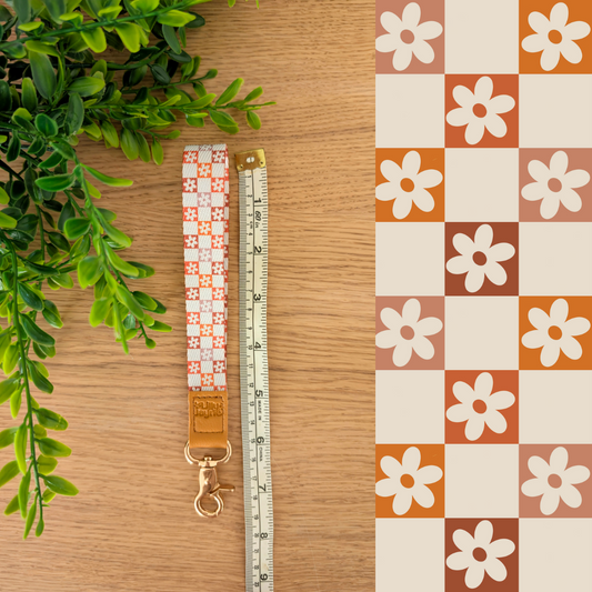Three colourful fabric wristlet keychains with gold hardware and tan leather ends, featuring pink gingham daisy, blush polka dot stripe, and retro orange checker floral patterns, laid on pastel notebooks with floral accents β Lilly Jayne Collective.