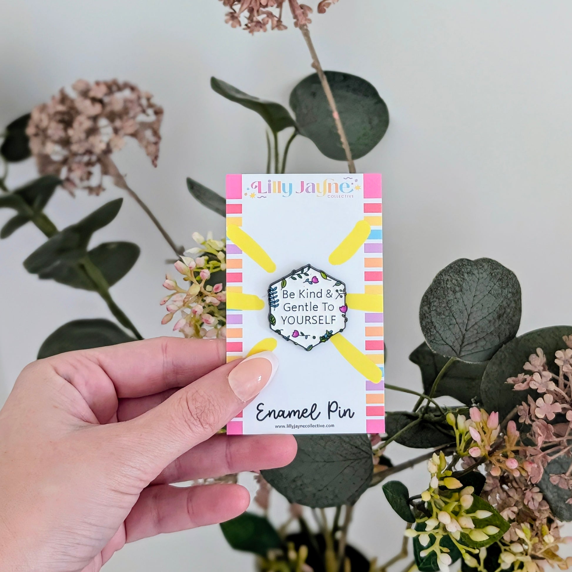 Hand holding a card with an enamel pin design, surrounded by plants.