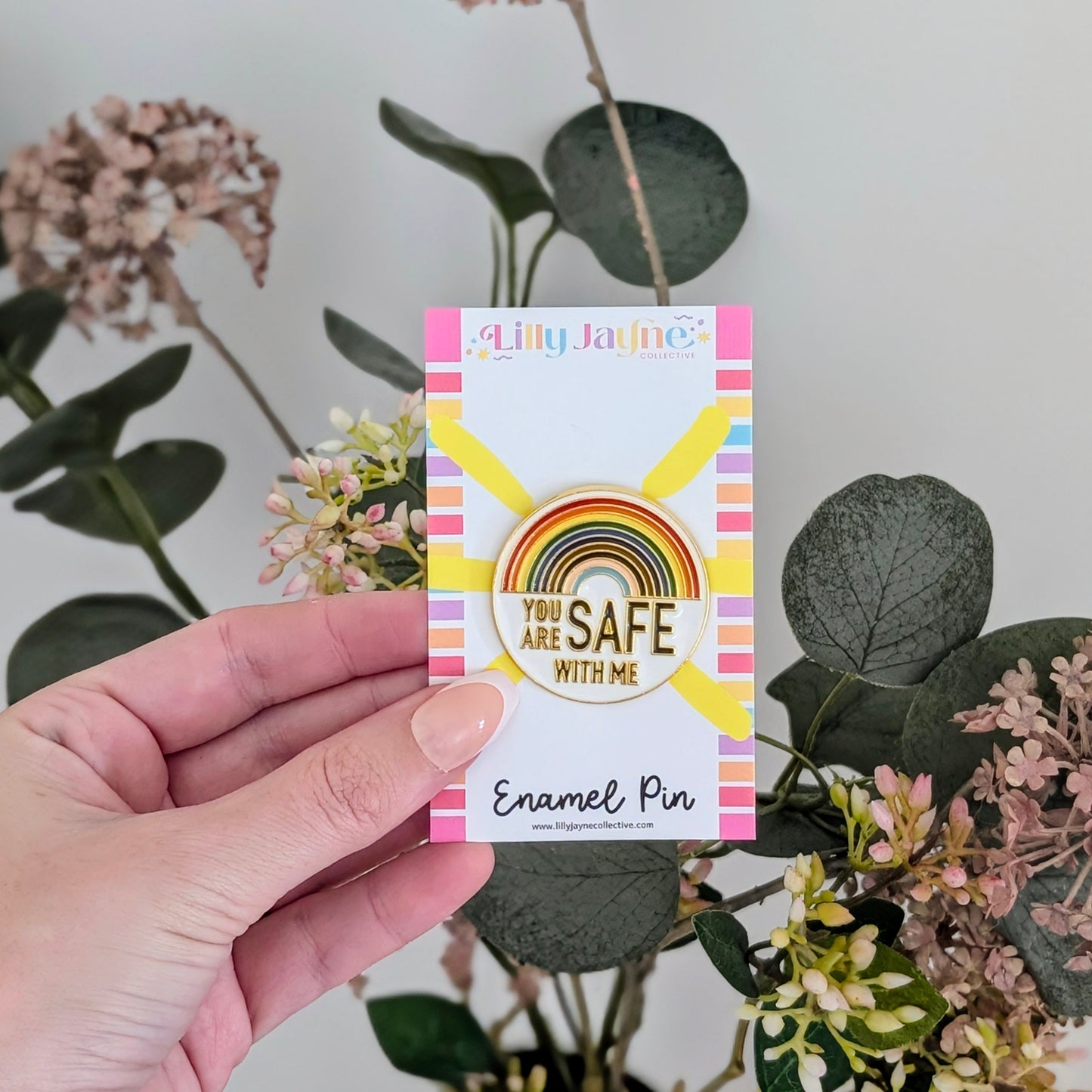 Hand holding a card with a rainbow-themed enamel pin and plants in the background