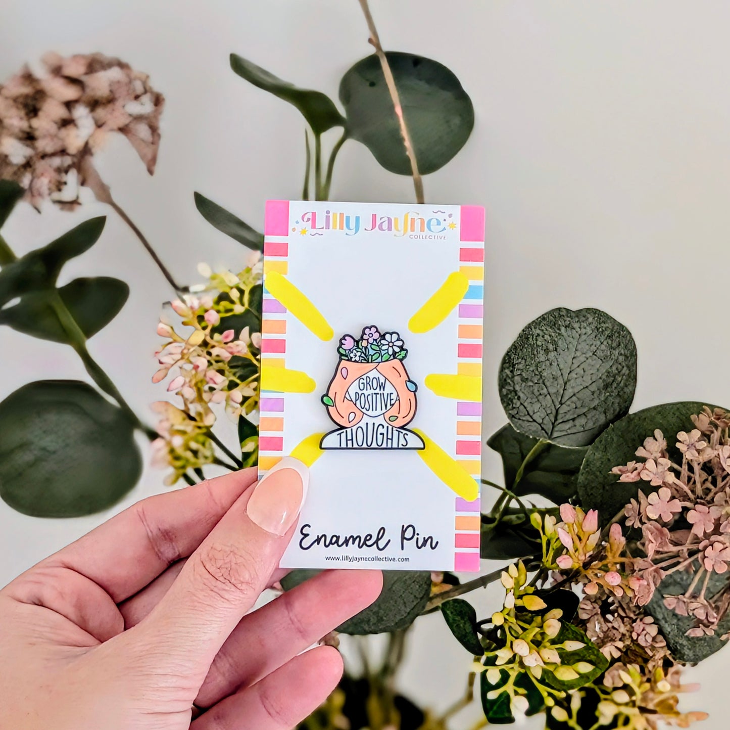 Hand holding a card with an enamel pin featuring a plant and text, surrounded by flowers and leaves.