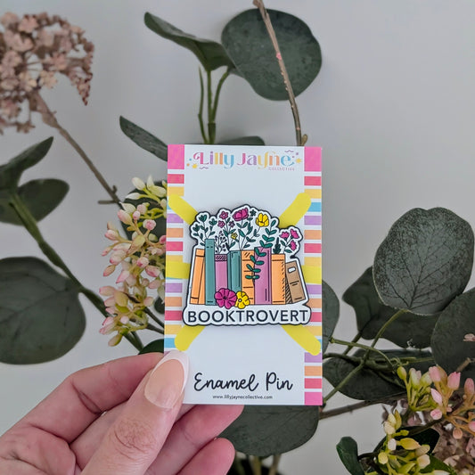 Enamel pin with 'Booktrovert' design on a card held in front of greenery