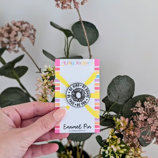 Hand holding a card with an enamel pin design, surrounded by flowers and plants.