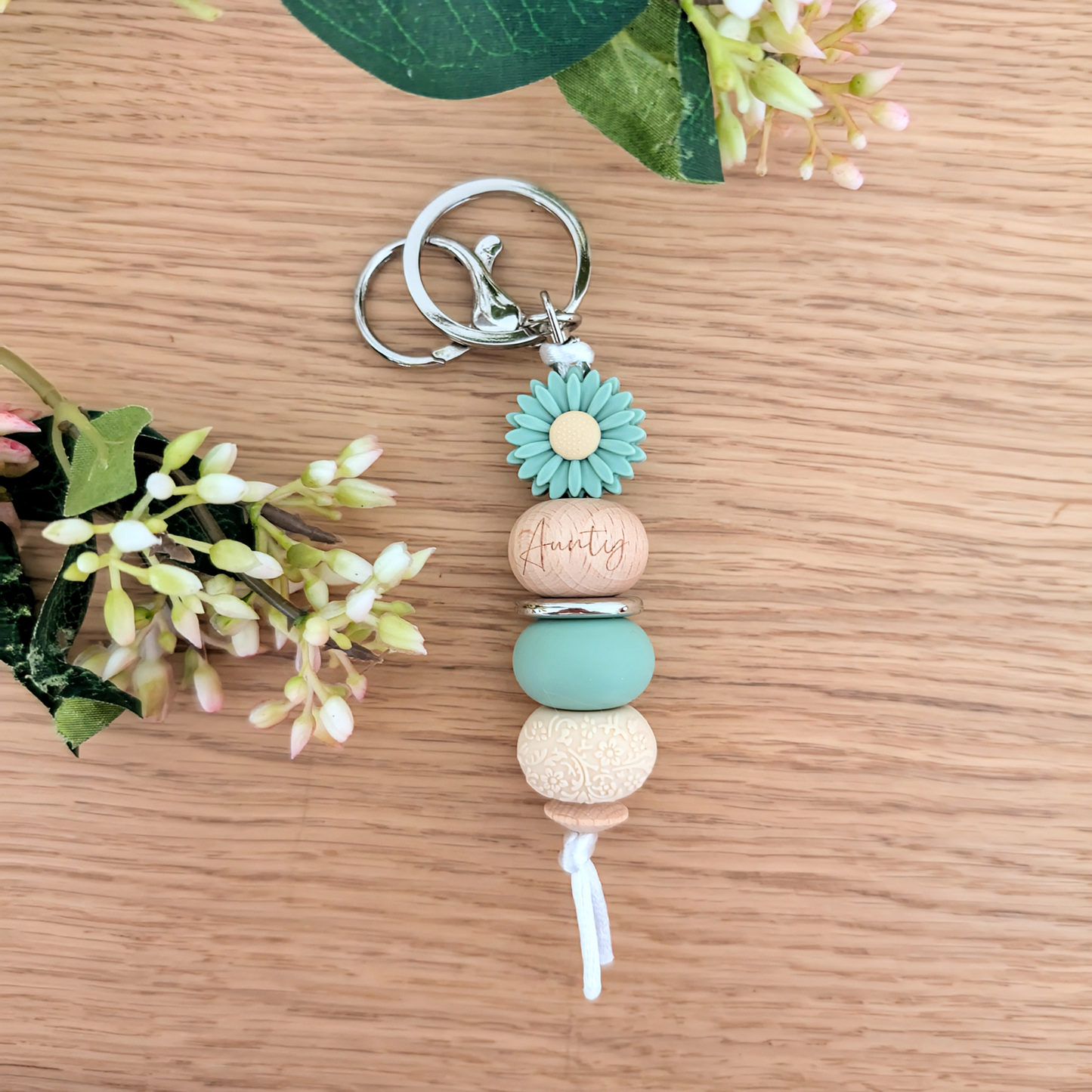 Handmade keyring with Sage Green daisy flower charm, engraved 'Aunty' wooden bead, floral patterned bead, and pastel pink accents on a wooden surface with greenery