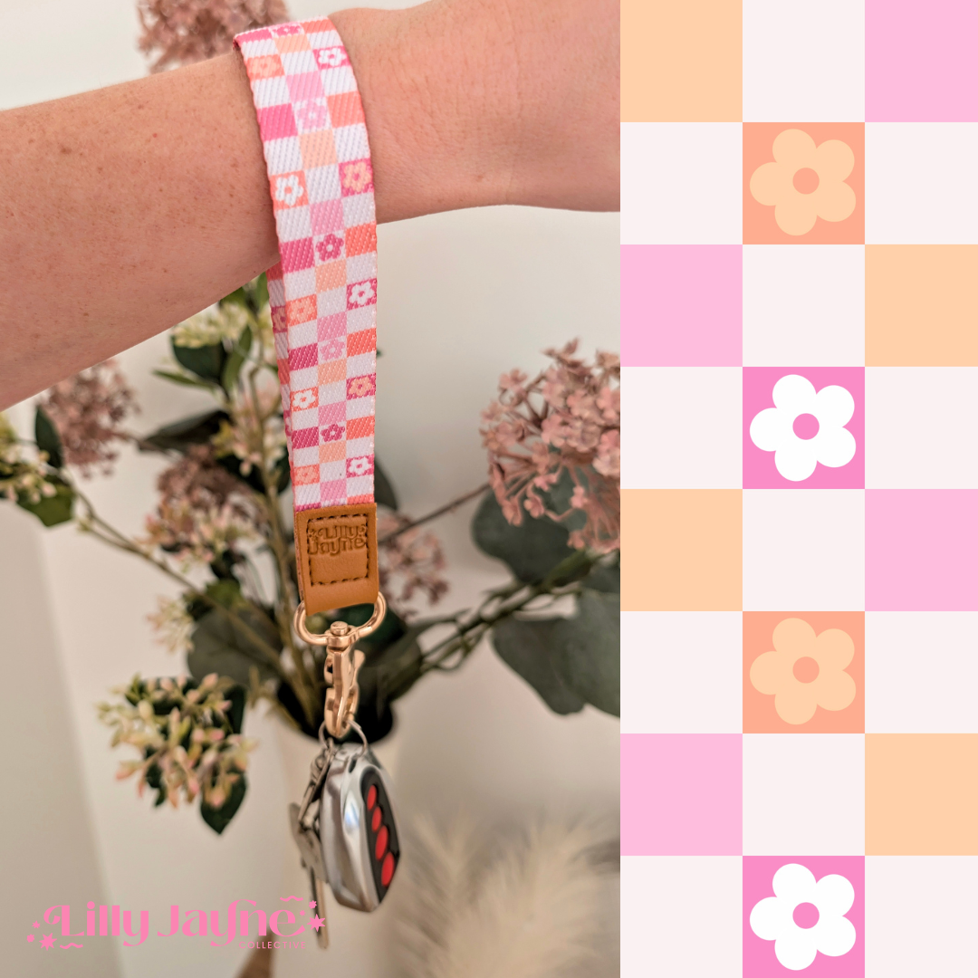 Three colourful fabric wristlet keychains with gold hardware and tan leather ends, featuring pink gingham daisy, blush polka dot stripe, and retro orange checker floral patterns, laid on pastel notebooks with floral accents β Lilly Jayne Collective.