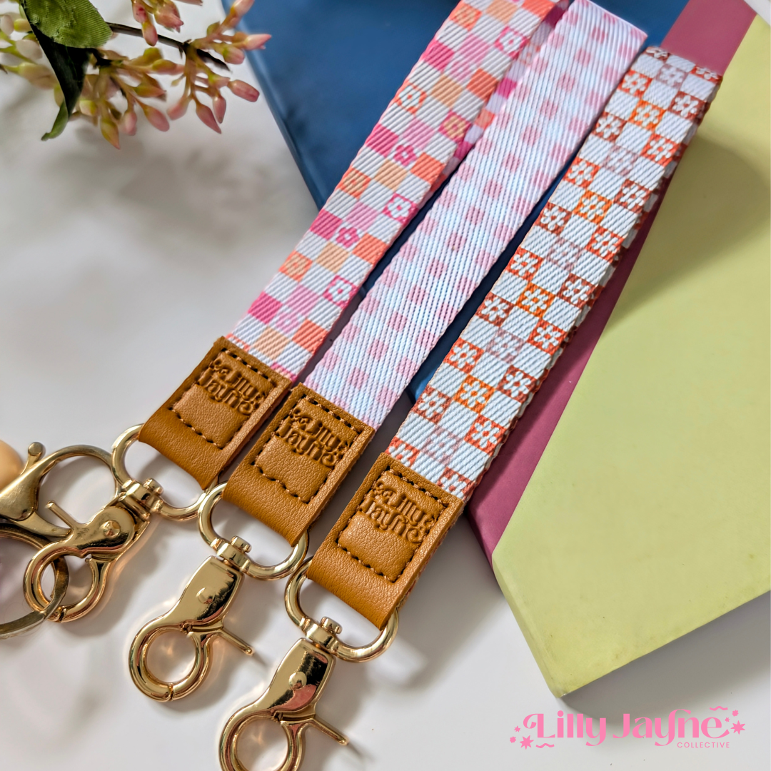 Three colourful fabric wristlet keychains with gold hardware and tan leather ends, featuring pink gingham daisy, blush polka dot stripe, and retro orange checker floral patterns, laid on pastel notebooks with floral accents β Lilly Jayne Collective.