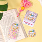 Colorful bookmarks and stickers on an open book with a decorative item on a yellow surface.
