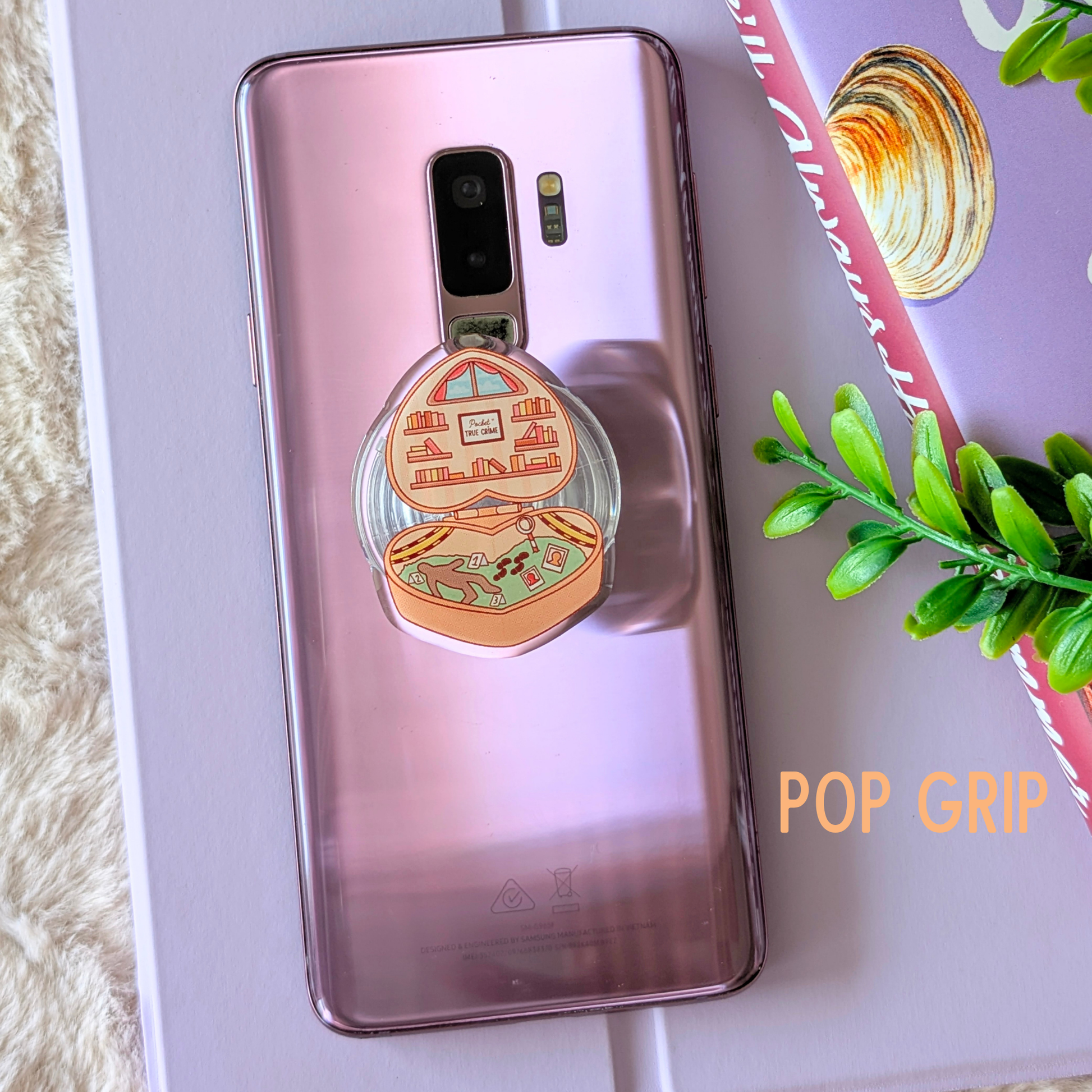 Phone case with a pop grip attachment on a pink phone, placed on a surface with a plant and magazine.