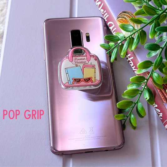 Phone case with pop grip attachment on a pink phone, next to a plant and a magazine.