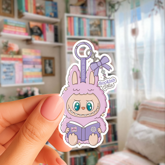 Keychain with cartoon character holding a book, held by a hand in a room with bookshelves.