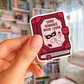 Sticker of a red book titled 'Dark Romance Book Lover' held by a hand in front of a bookshelf.