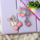 Pink cherry and bow-themed phone holder on a light purple surface with green leaves in the background.