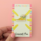Enamel pin on a colorful card held by a hand against a pink background
