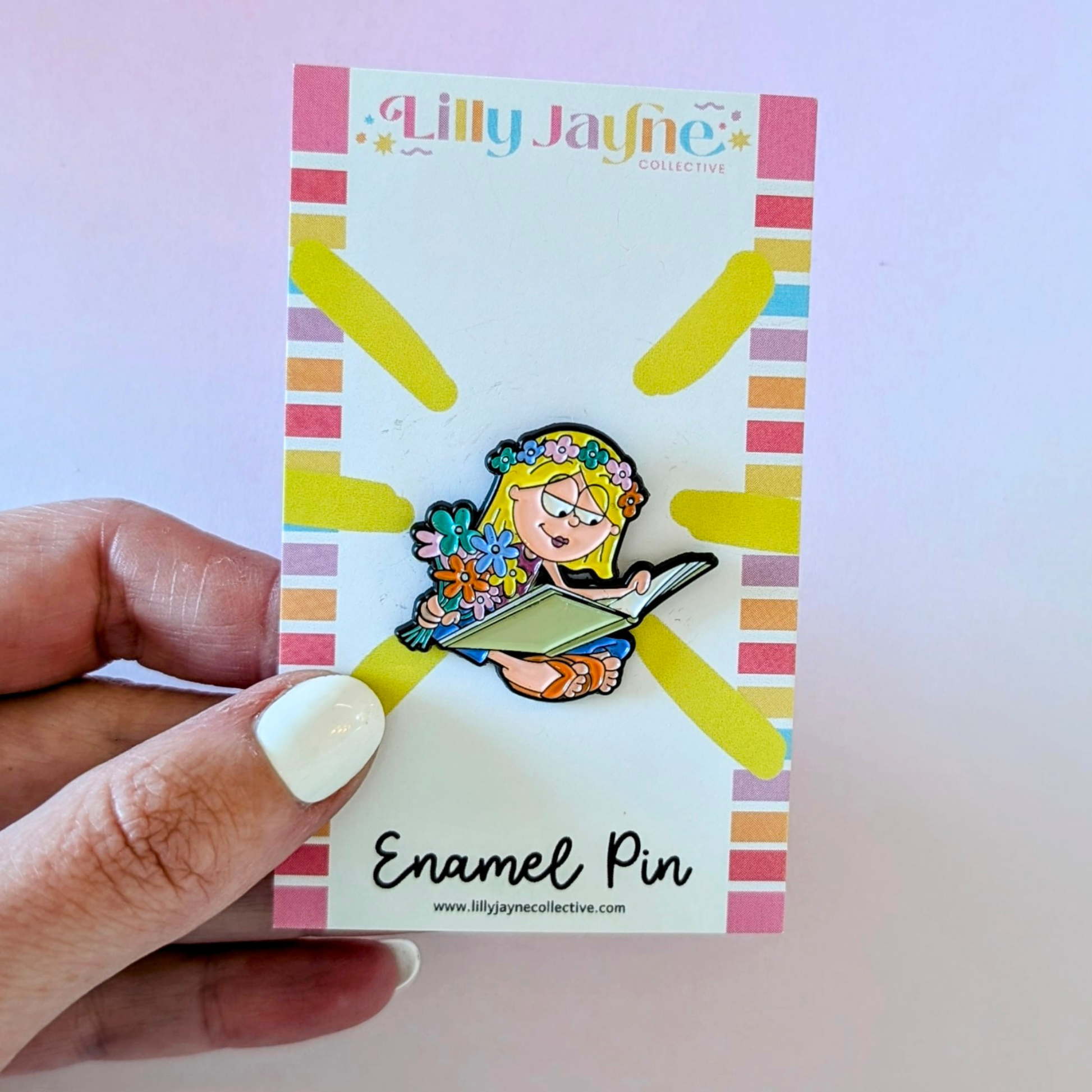 Enamel pin on a colorful card held by a hand with white nail polish.