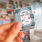 Sticker of a book titled 'Thriller Book Lover' held by a hand with a blurred bookshelf background.