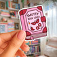 Sticker of a pink book titled 'Smutty Book Lover' held by a hand with a blurred bookshelf background.