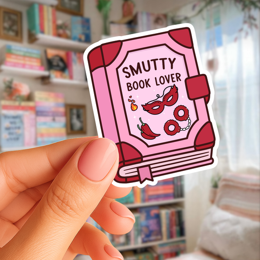 Sticker of a pink book titled 'Smutty Book Lover' held by a hand with a blurred bookshelf background.