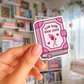 Pink book-shaped sticker with 'Slow Burn Book Lover' text held in front of a bookshelf.