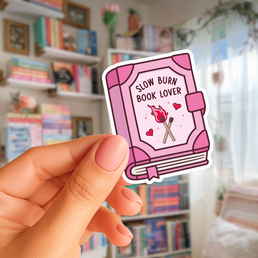 Pink book-shaped sticker with 'Slow Burn Book Lover' text held in front of a bookshelf.