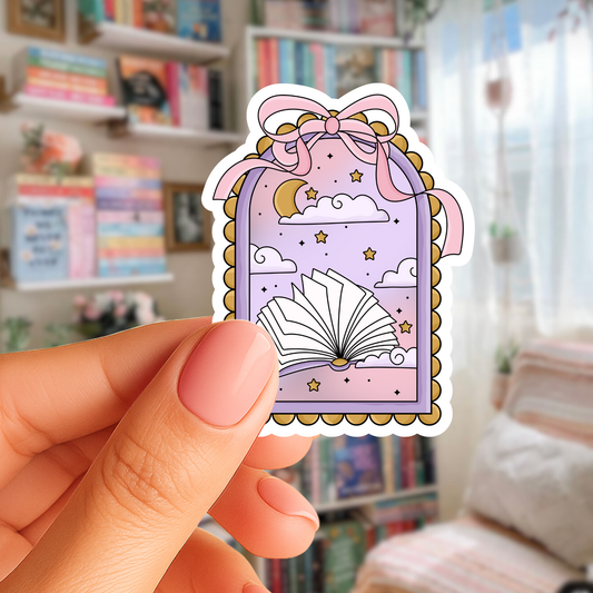 Hand holding a decorative sticker with a purple frame, book, and clouds against a blurred indoor background.