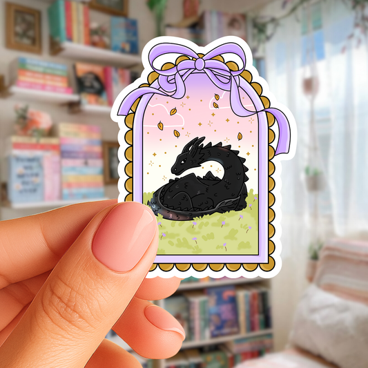 Hand holding a sticker of a black dragon in a decorative frame with a bow against a blurred indoor background.