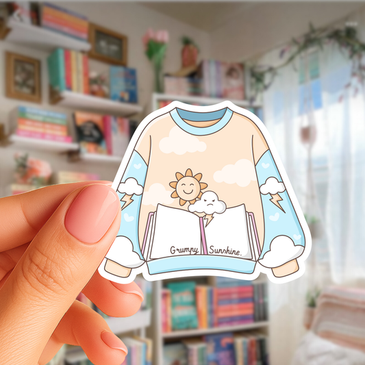 Hand holding a sticker of a sweater with a sun and clouds design in a room with bookshelves.