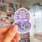Hand holding a colorful sticker of a fantasy scene with books and a castle in a room with bookshelves.