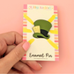 Enamel pin on a card held by a hand against a pink background