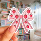 Sticker of a pink bow with strawberry patterns held by a hand in a room with books and decor.