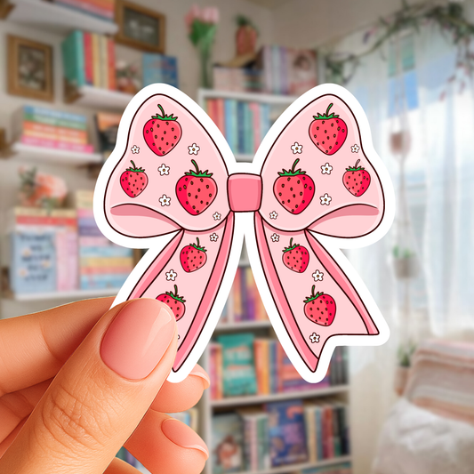 Sticker of a pink bow with strawberry patterns held by a hand in a room with books and decor.