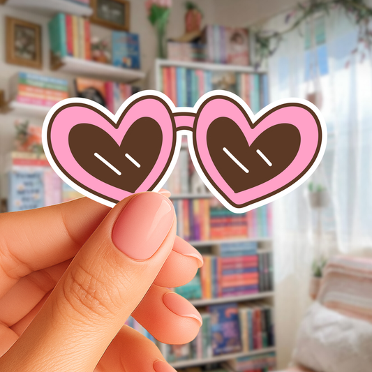 Heart-shaped sunglasses held by a hand in front of a bookshelf