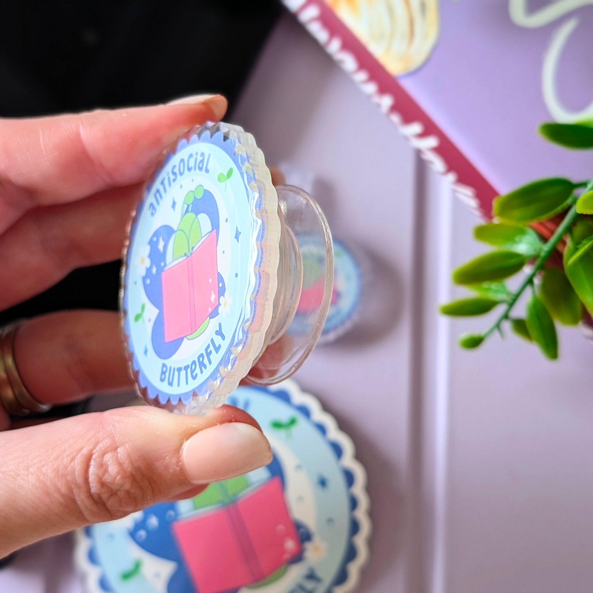Hand holding a colorful sticker with 'antisocial butterfly' text against a blurred background