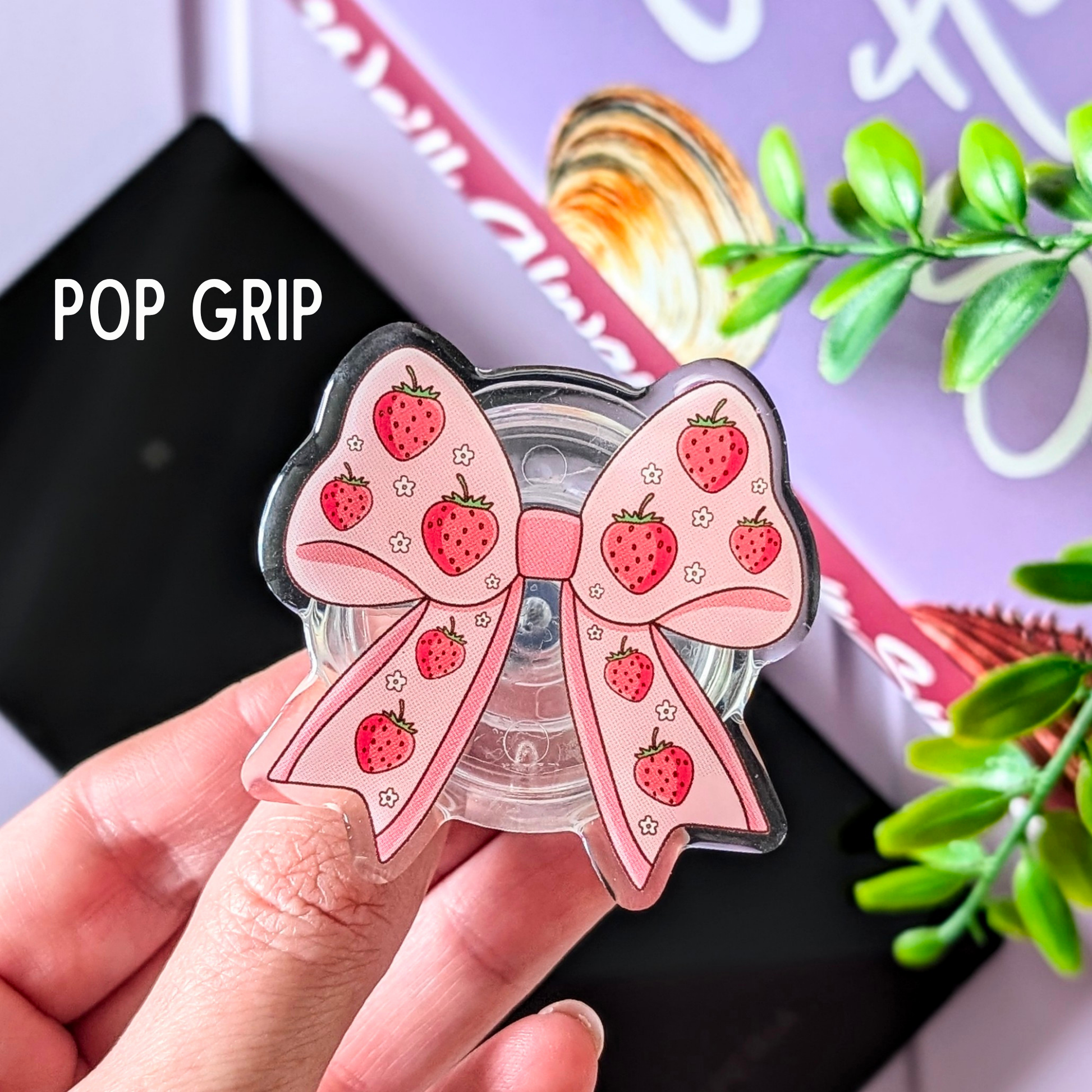 Pink bow-shaped pop grip with strawberry design held by a hand, with a blurred background.
