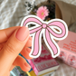 Hand holding a pink bow-shaped sticker with a blurred background of books and flowers.