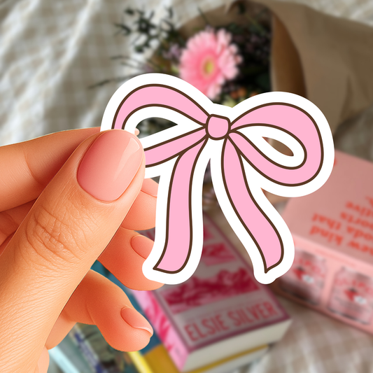 Hand holding a pink bow-shaped sticker with a blurred background of books and flowers.
