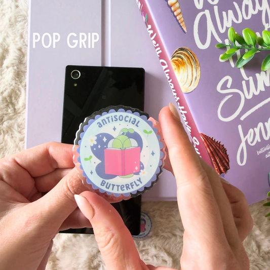 Hand holding a round pop grip with a design of a butterfly and book, against a blurred background with a phone and book.