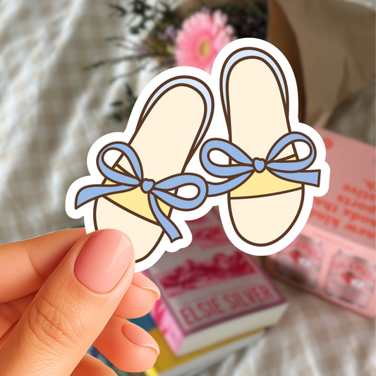 Hand holding a sticker of ballet shoes with blue bows against a blurred background