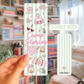 Double-sided bookmark with 'Camp Stories' design held by a hand in front of a bookshelf.