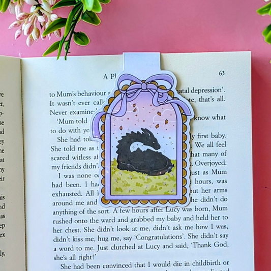 Decorative bookmark with a dragon illustration on an open book against a pink background.