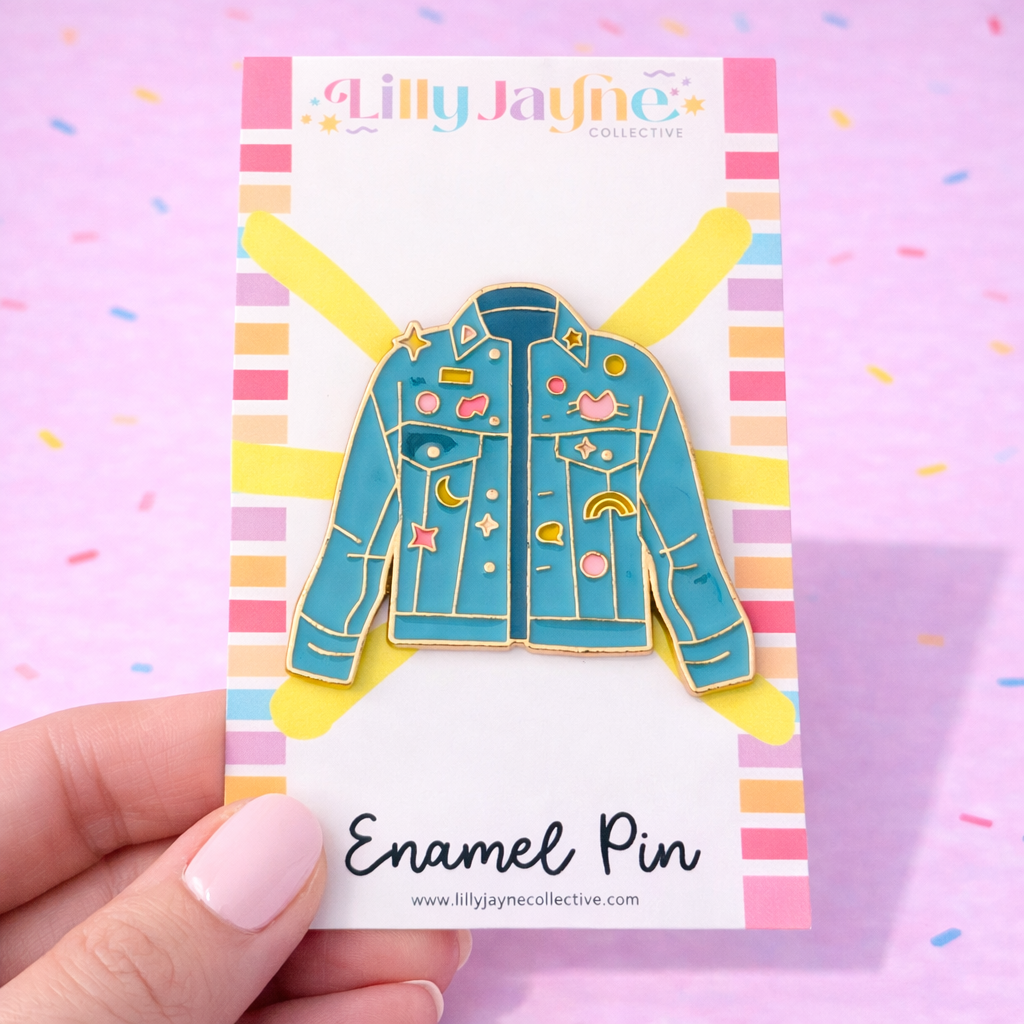 Enamel pin of a blue jacket with colorful details on a card held by a hand against a colorful background.