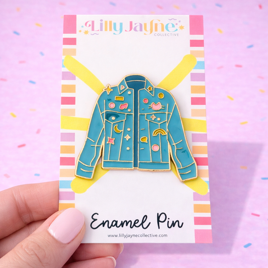 Enamel pin of a blue jacket with colorful details on a card held by a hand against a colorful background.