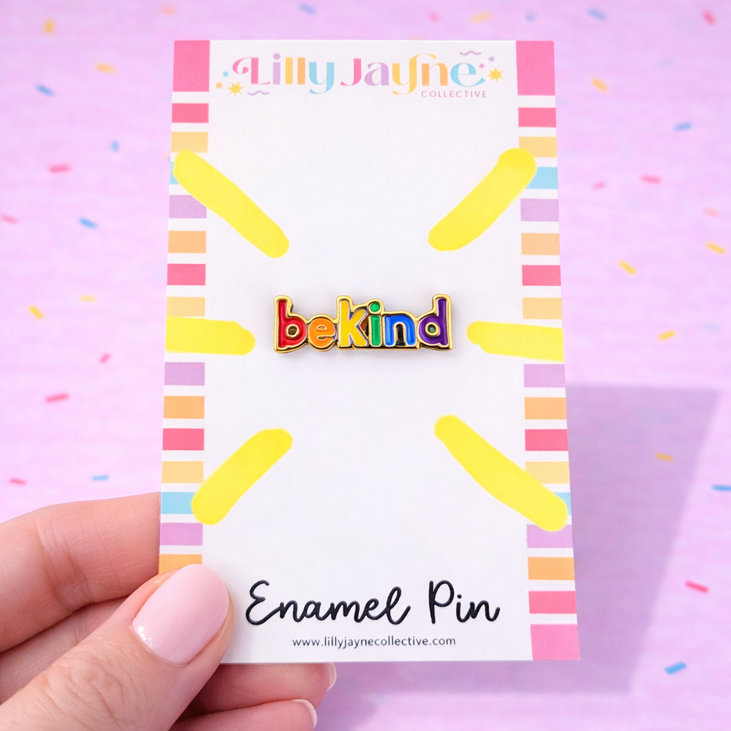 Enamel pin with 'bekind' text on a colorful card held by a hand against a pink background.