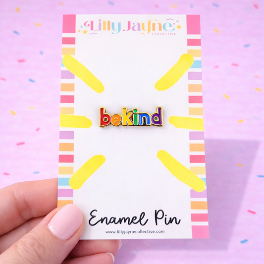 Enamel pin with 'bekind' text on a colorful card held by a hand against a pink background.